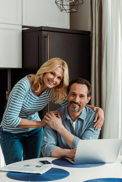 Selektiver Fokus einer reifen Frau, die in die Kamera lächelt, während sie ihren Mann in der Nähe digitaler Geräte auf dem Küchentisch umarmt  - Foto, Bild