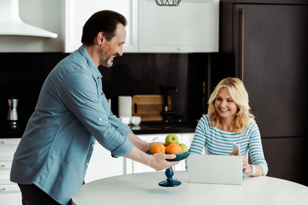 Smiling man holding vase with fruits near wife using smartphone and laptop in kitchen  - Foto, Imagem
