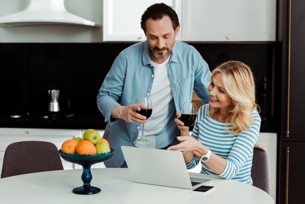 Smiling woman holding glass of wine and pointing on laptop near husband in kitchen   - Foto, imagen