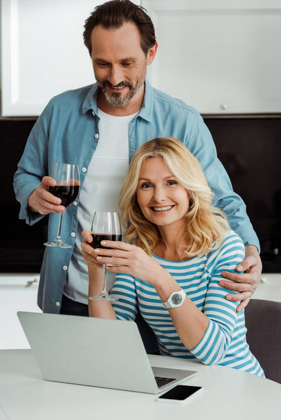 Schöner Mann umarmt lächelnde Frau mit Glas Wein in der Nähe von Gadgets auf Küchentisch  - Foto, Bild