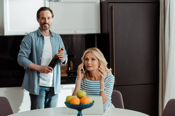 Selectieve focus van lachende man met fles wijn terwijl vrouw praten op smartphone in de buurt van laptop in de keuken  - Foto, afbeelding