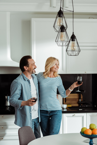 Selective focus of smiling man embracing wife with glass of wine in kitchen  - Photo, Image