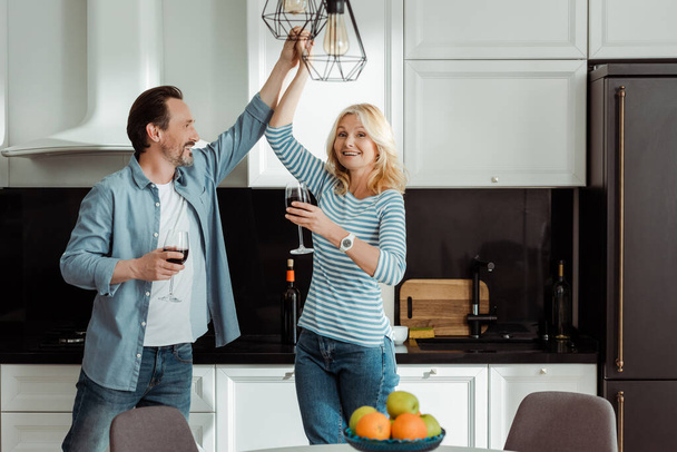 Selective focus of smiling mature couple dancing with glasses of wine in kitchen  - Photo, Image