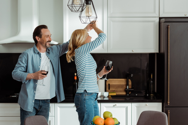 Selective focus of cheerful mature couple dancing with wine glasses in kitchen  - Φωτογραφία, εικόνα
