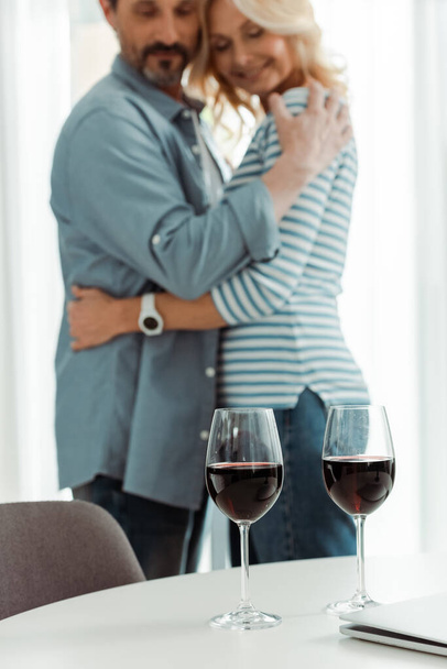 Selektywne skupienie kieliszków wina i laptopa na stole w pobliżu uśmiechniętej dojrzałej pary obejmującej kuchnię  - Zdjęcie, obraz