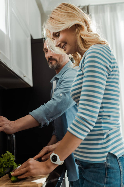 Side view of smiling woman cutting cucumber near husband in kitchen  - Photo, Image