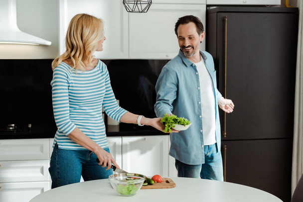 Smiling man giving lettuce to wife cutting vegetables in kitchen  - Photo, Image