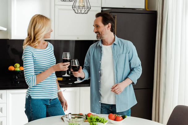 Lächelndes älteres Paar klingelt mit Wein neben frischem Gemüse auf dem Küchentisch  - Foto, Bild