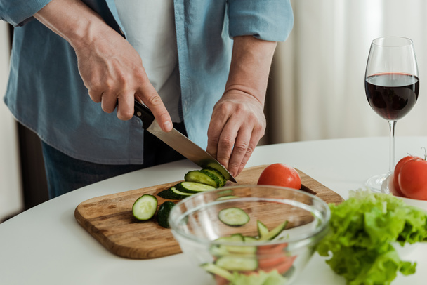 Cropped view of man cutting cucumber while cooking salad near glass of wine in kitchen  - Photo, image
