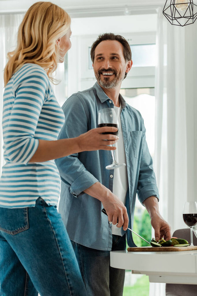 Selective focus of man smiling at wife with glass of wine during cooking salad in kitchen  - Foto, immagini