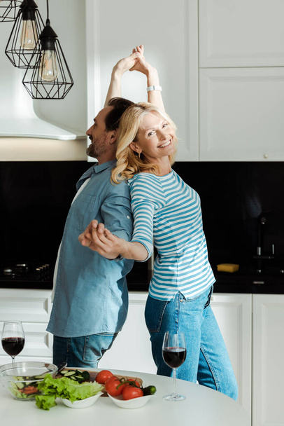 Side view of smiling mature couple dancing near fresh salad and wine on kitchen table  - Photo, Image