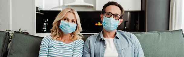 Panoramic shot of mature couple in medical masks sitting on couch at home  - Zdjęcie, obraz