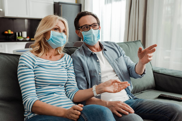 Enfoque selectivo de la mujer en la máscara médica viendo la televisión cerca de marido señalando con la mano en el sofá
  - Foto, Imagen