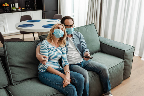 Mature man hugging wife in medical mask while watching movie on couch  - Фото, изображение