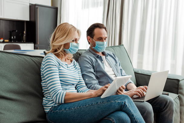 Reifes Paar in medizinischen Masken mit Laptop und digitalem Tablet auf der Couch  - Foto, Bild
