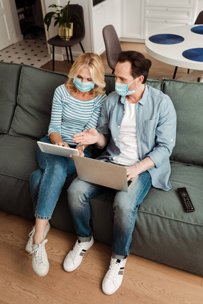 ノートパソコンを使用した医療用マスクの男性の高い角度のビューとソファの上にデジタルタブレットを使用して妻の近くの手で指す  - 写真・画像