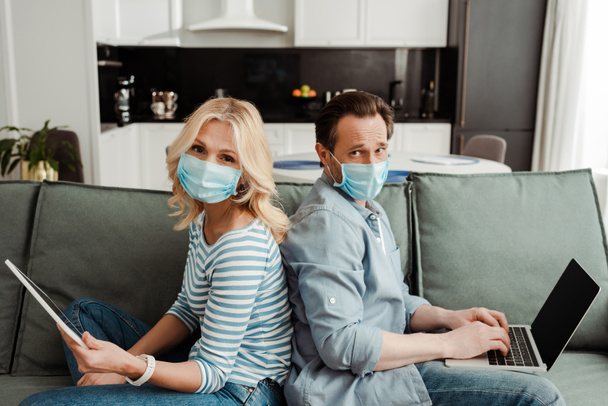 Vue latérale du couple mature dans des masques médicaux regardant la caméra tout en utilisant des gadgets sur le canapé
  - Photo, image