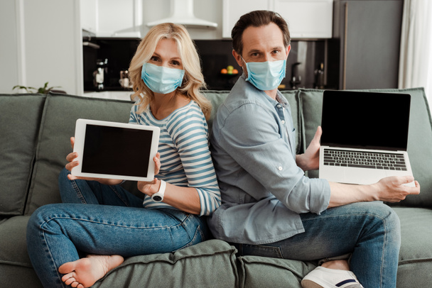 Widok z boku dojrzałej pary w maskach medycznych pokazujących laptopa i tabletu cyfrowego z pustymi ekranami na kanapie  - Zdjęcie, obraz