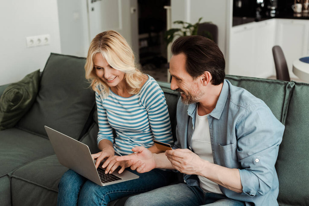 Handsome man pointing with finger while holding credit card near smiling woman using laptop at home  - Photo, Image