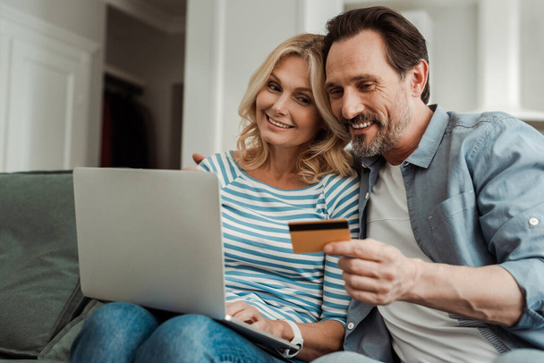 Επιλεκτική εστίαση του χαμογελαστού ώριμου ζευγαριού χρησιμοποιώντας πιστωτική κάρτα και φορητό υπολογιστή στον καναπέ  - Φωτογραφία, εικόνα
