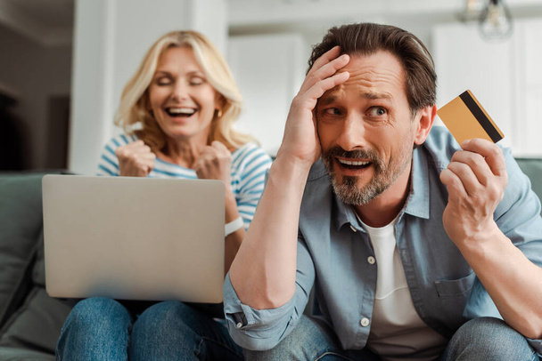 Confused man holding credit card near cheerful wife using laptop at home  - Photo, Image