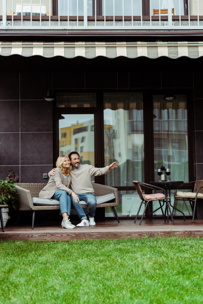 Reifer Mann zeigt mit der Hand und umarmt lächelnde Frau auf Weidensofa auf der Terrasse - Foto, Bild