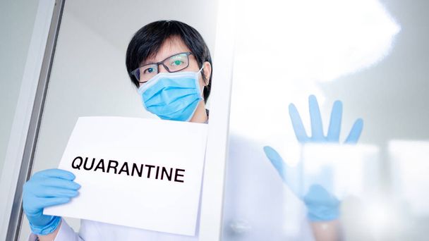Мужчина азиатский врач или врач мужчина в хирургической маске и перчатках, показывающих бумагу с сообщением Оставайтесь дома в клинике больницы. Карантинная кампания во время пандемии коронавируса (COVID-19)
. - Фото, изображение