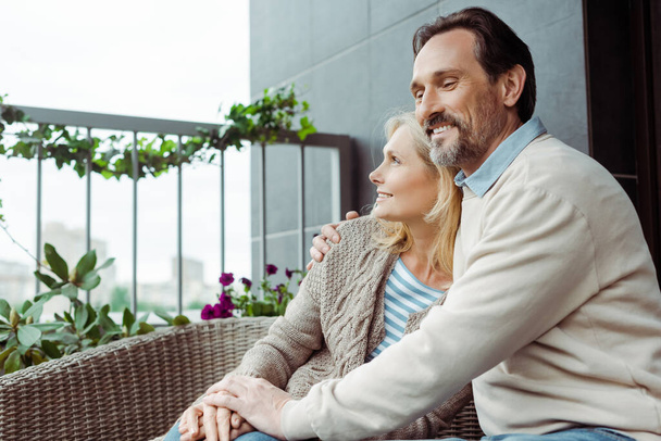 Mature man hugging beautiful smiling wife on sofa on terrace  - Фото, изображение