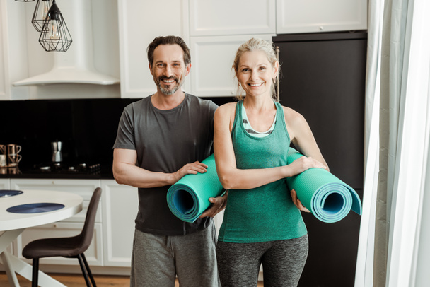 volwassen paar in sportkleding glimlachen op camera terwijl het houden van fitness matten thuis  - Foto, afbeelding