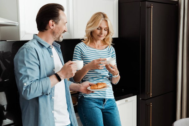 Улыбающийся мужчина держит круассан рядом с женой с чашкой кофе на кухне
  - Фото, изображение