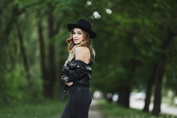 Portrait of a model girl in leather jacket and black hat posing alone on a summer city street - 写真・画像