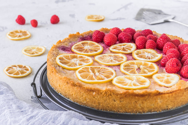 Cheese cake with raspberries and dried lemon slices on white background, some raspberries, lemon slices and cake server lying on the table. Closeup. - Photo, Image