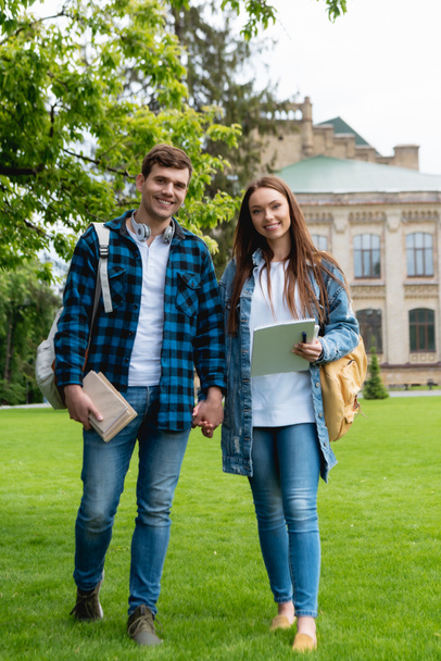 cheerful students holding hands while standing on grass near university campus  - Fotoğraf, Görsel