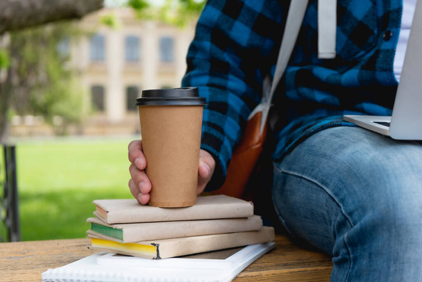 καλλιεργημένη άποψη του μαθητή κρατώντας χάρτινο κύπελλο κοντά σε βιβλία - Φωτογραφία, εικόνα