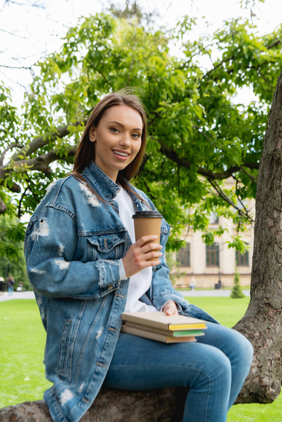 cheerful and beautiful student holding paper cup and books while sitting on tree  - Fotó, kép