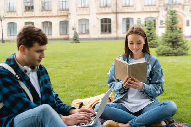 selektiver Fokus fröhlicher Mädchen beim Lesen von Büchern in der Nähe gutaussehender Schüler mit Laptop, Online-Studienkonzept  - Foto, Bild
