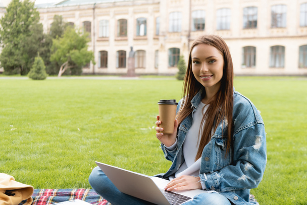 estudante atraente segurando copo descartável e sorrindo ao usar laptop, conceito de estudo on-line
  - Foto, Imagem