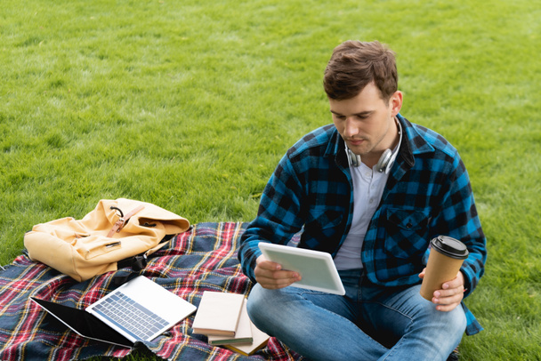 handsome student holding digital tablet and paper cup near laptop and books on plaid blanket, online study concept  - Foto, Imagem