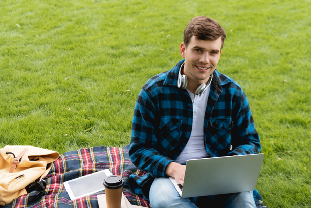 happy student using laptop near digital tablet and paper cup on plaid blanket, online study concept  - Foto, Bild