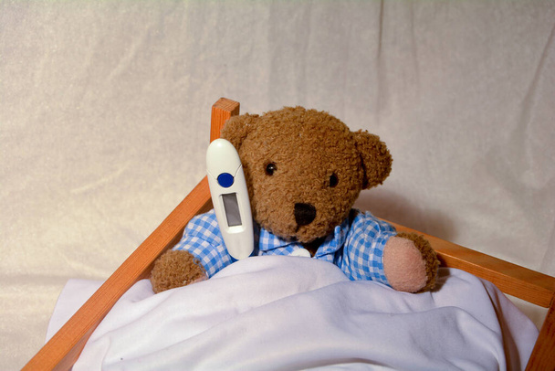 Um ursinho doente está sentado na cama com um termômetro clínico
 - Foto, Imagem