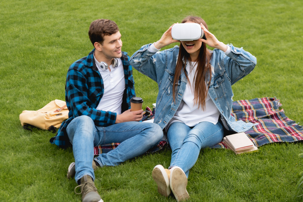 estudante alegre segurando copo de papel e olhando para menina animado em fone de ouvido realidade virtual
  - Foto, Imagem