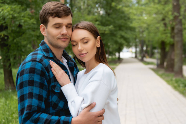 handsome boyfriend and pretty girlfriend with closed eyes hugging in park  - 写真・画像