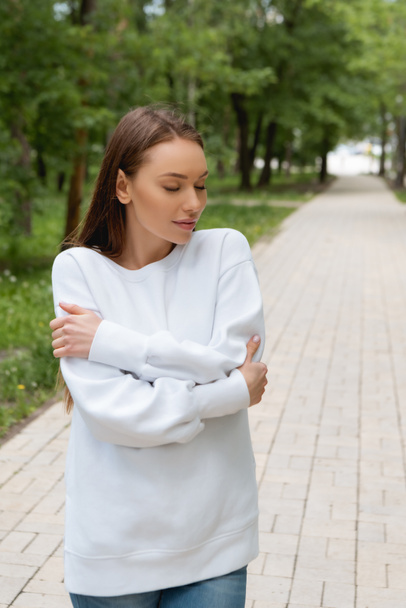 attractive young woman in sweatshirt standing with crossed arms in park  - Foto, afbeelding