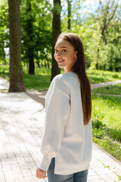 fröhliches Mädchen in weißem Sweatshirt steht im Park und blickt in die Kamera  - Foto, Bild