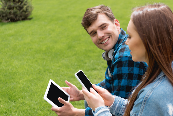 Selektiver Fokus des fröhlichen Schülers, der Mädchen mit Smartphone anschaut, während er ein digitales Tablet mit leerem Bildschirm hält - Foto, Bild