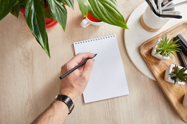 partial view of man writing in notepad near green plants on wooden surface - Photo, Image