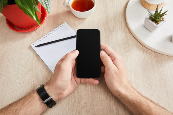 cropped view of man holding smartphone near green plants, cup of tea and blank notebook with pencil on wooden surface - Foto, imagen
