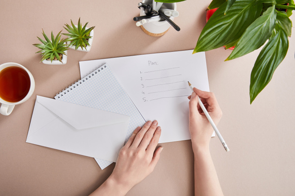 περικοπή άποψη της γυναίκας γραπτώς σχέδιο σε χαρτί κοντά σε πράσινα φυτά, φλιτζάνι τσάι, φάκελο, κενό σημειωματάριο σε μπεζ επιφάνεια - Φωτογραφία, εικόνα