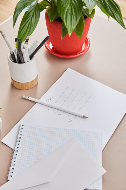 зеленое растение, конверт, пустой блокнот, карандаши и ручки и бумага с плановыми надписями на бежевой поверхности
 - Фото, изображение