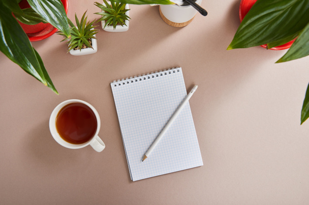 top view of green plants, cup of tea, blank notebook with pencil on beige surface - Fotoğraf, Görsel